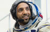 Emirati astronaut Sultan Al Neyadi with grey white space suit with UAE flag, helmet removed. He sits in front of the Soyuz spacecraft simulator in 2019.